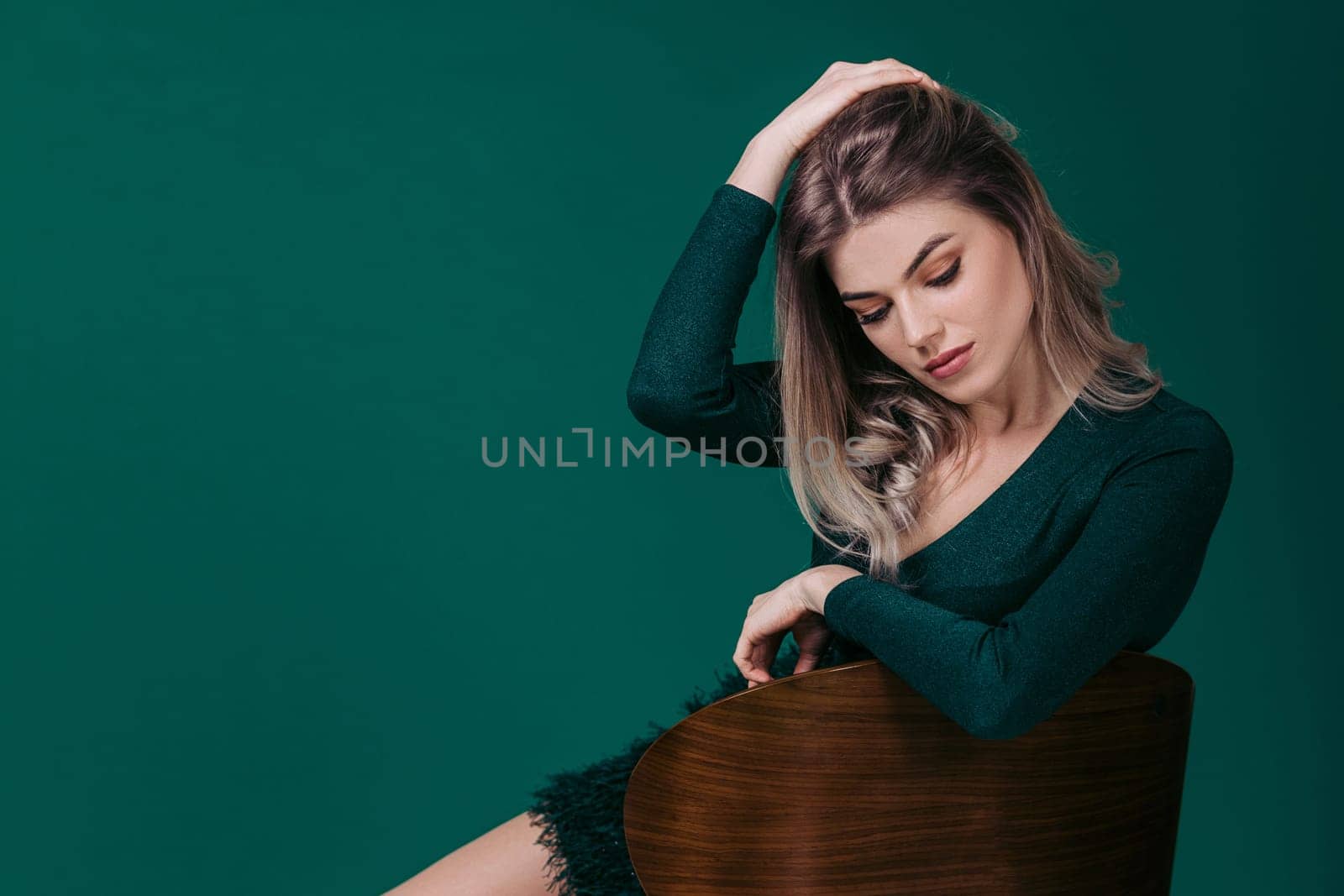 Sensual beautiful blonde woman in green dress sitting on chair against green background