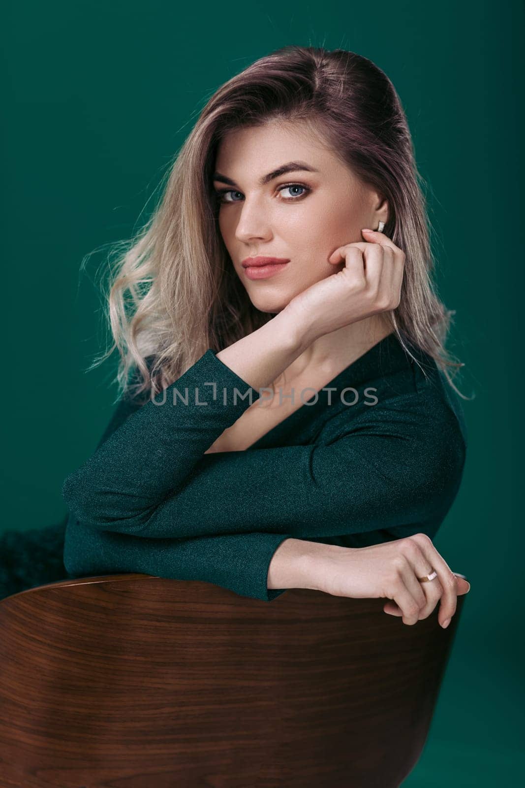 Sensual beautiful blonde woman in green dress sitting on chair and looking at camera against green background