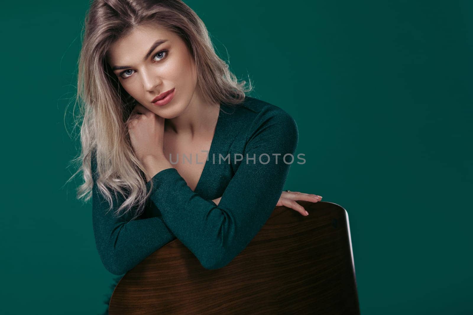 fashion portrait of sensual beautiful blonde woman in green dress sitting on chair and looking at camera against green background