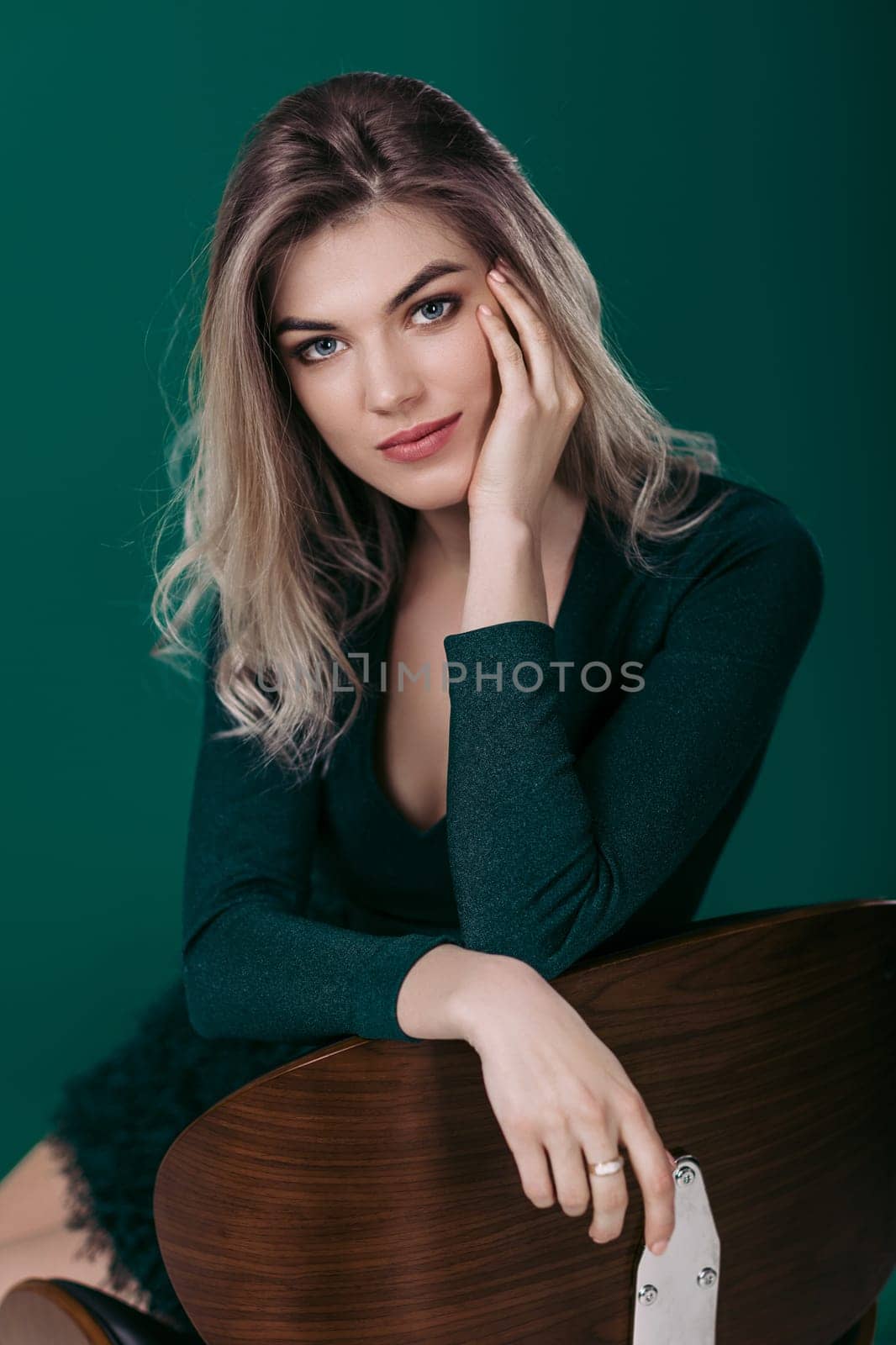Sensual beautiful blonde woman in green dress sitting on chair and looking at camera against green background