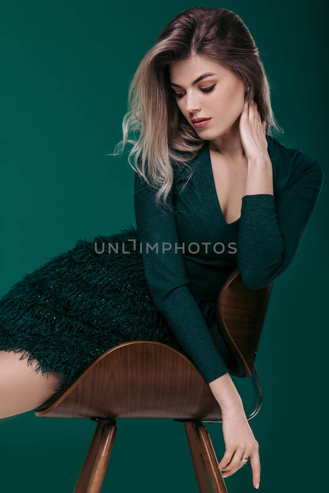 Sensual beautiful blonde woman in green dress sitting on chair against green background