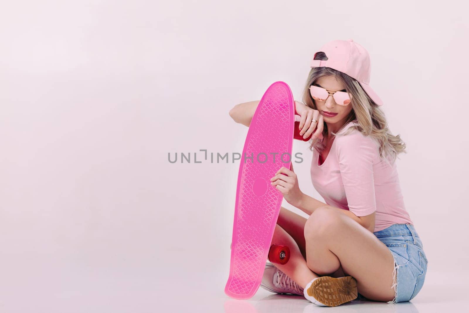 Cute woman in sunglasses, pink t-shirt and cap sitting on the floor with pink skateboard. teenager