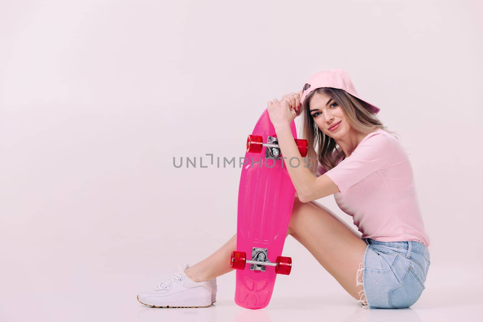 Cute woman in pink t-shirt and cap sitting on the floor with pink skateboard. teenager