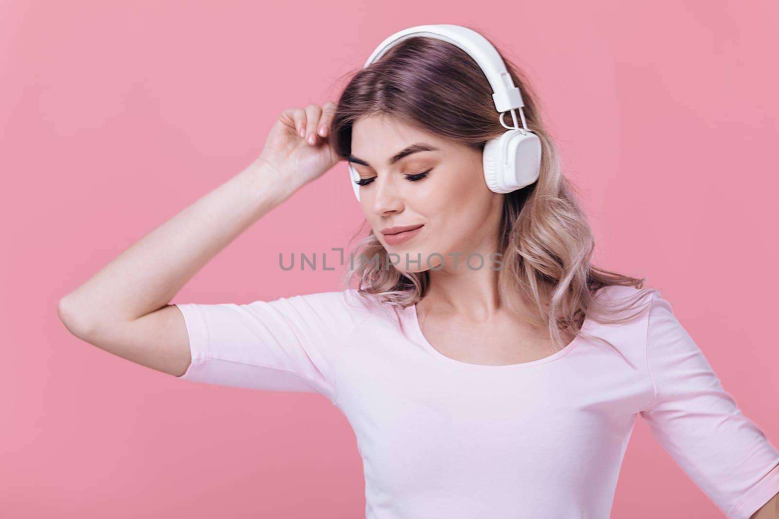 beautiful blonde woman in pink t-shirt and white headphones listens to music and smiling with closed eyes on pink background