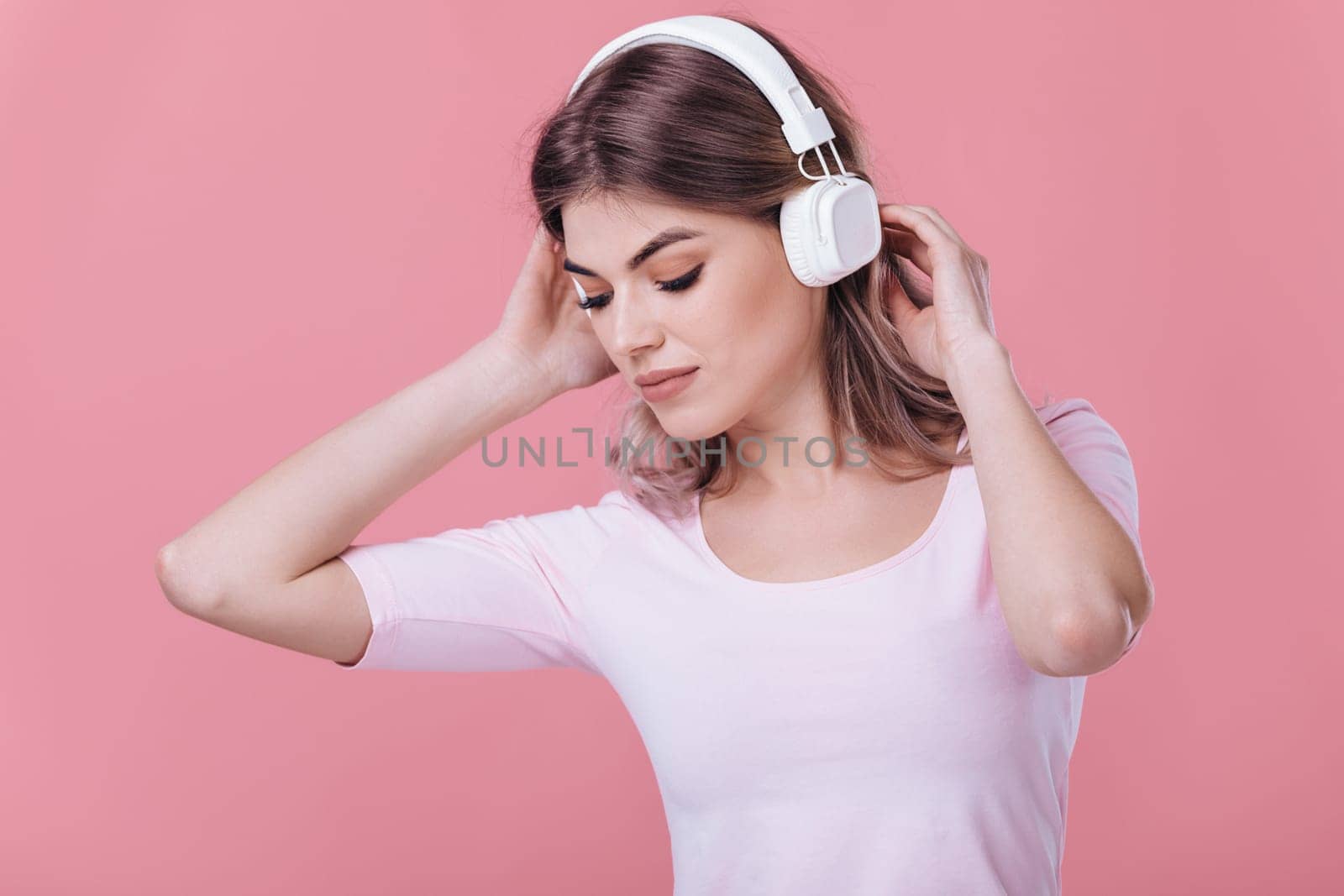 beautiful blonde woman in pink t-shirt and white headphones listens to music on pink background