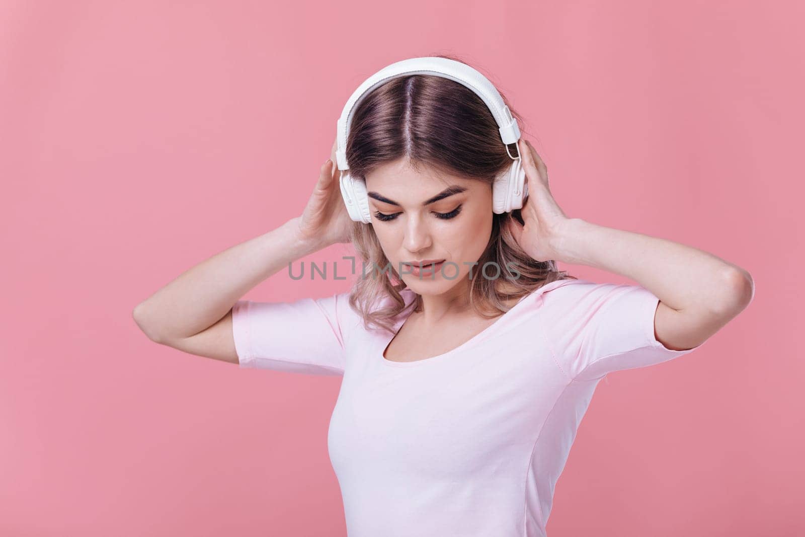 beautiful blonde woman in pink t-shirt and white headphones listens to music and smiling with closed eyes on pink background