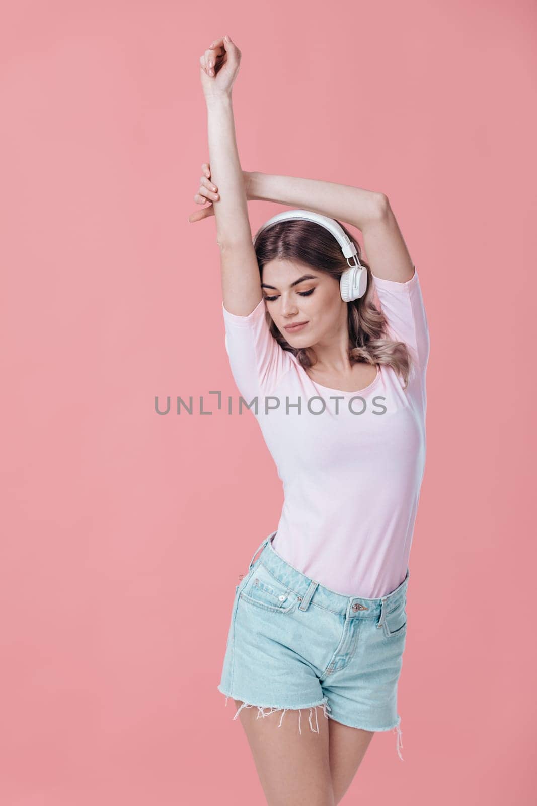 beautiful blonde woman in pink t-shirt and white headphones listens to music on pink background. summer studio photoshoot