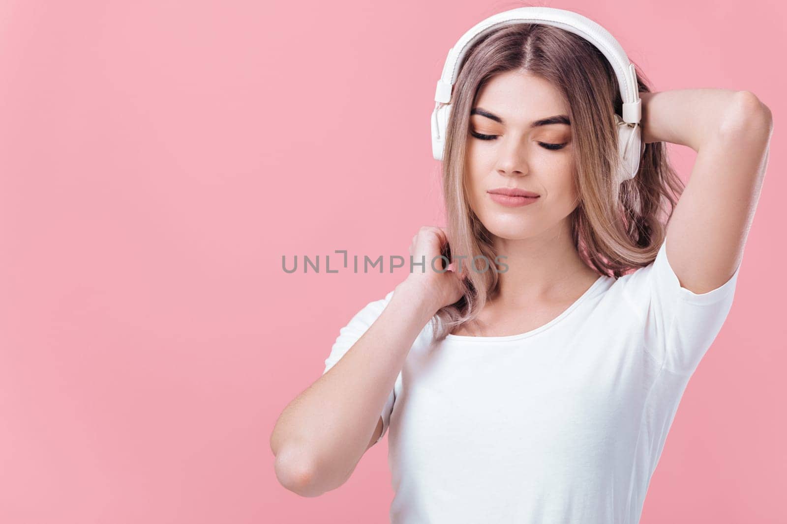 beautiful blonde woman in t-shirt and white headphones enjoying listens to music on pink background