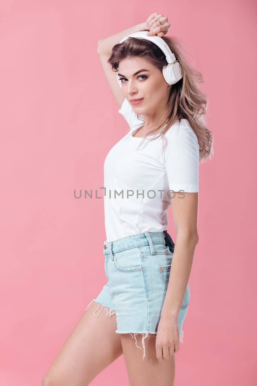 beautiful blonde woman in t-shirt and white headphones listens to music on pink background