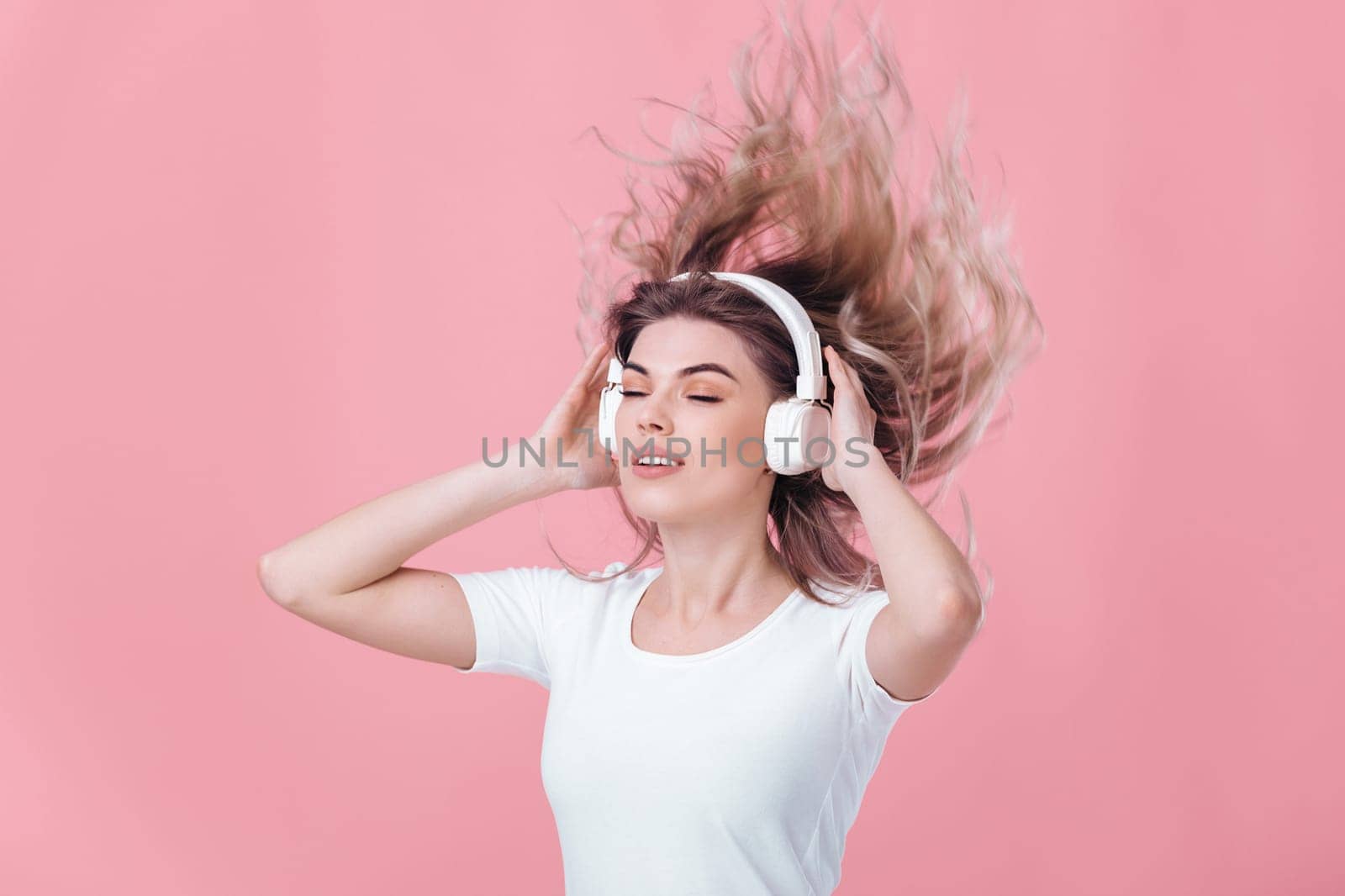 beautiful blonde woman in t-shirt and white headphones listens to music on pink background. hair fly