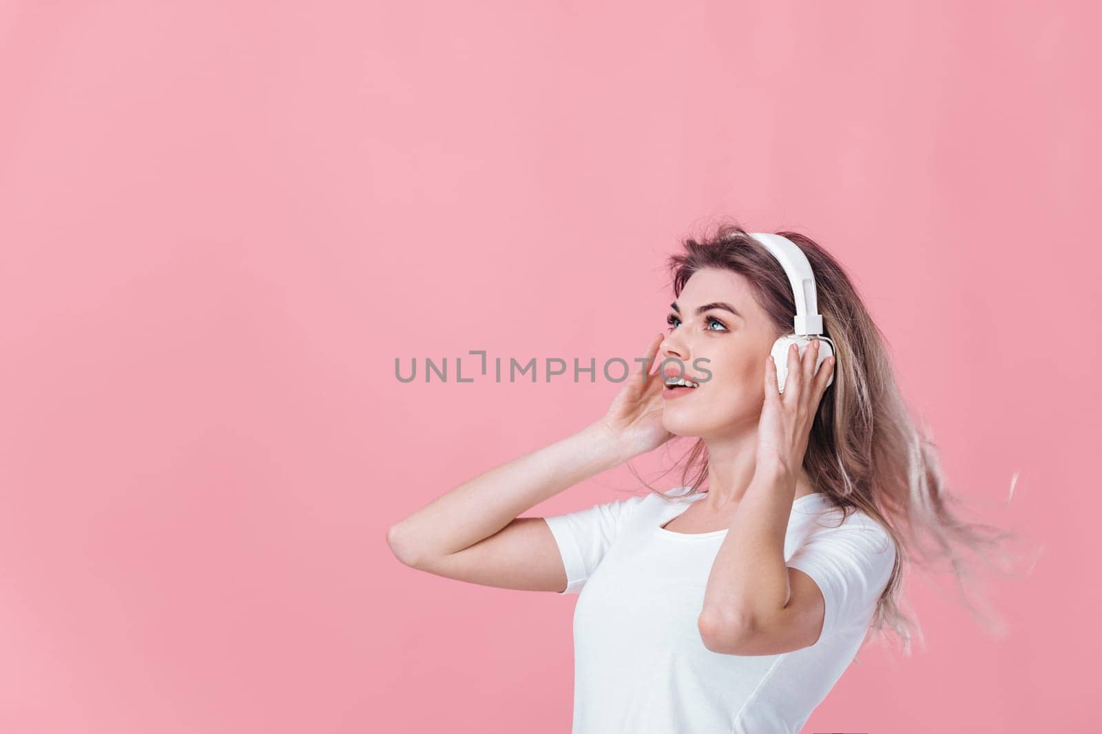 beautiful smiling blonde woman in t-shirt and white headphones listens to music on pink background