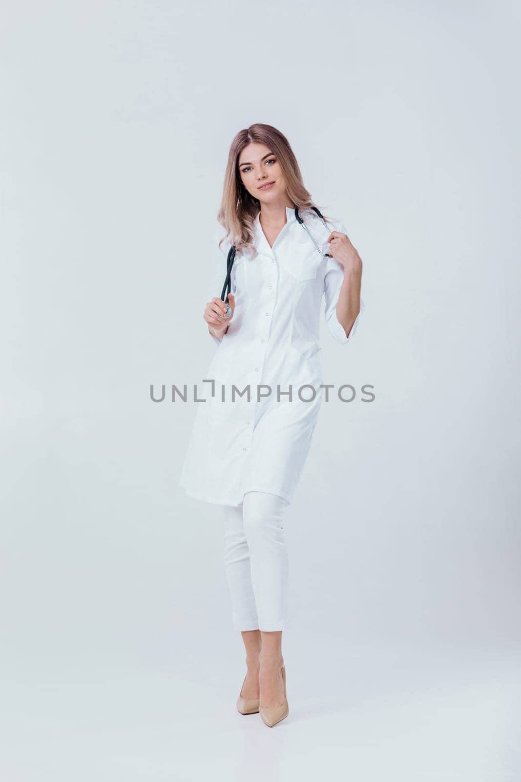 Full length portrait of medical physician doctor woman in white coat with stethoscope looks at camera on light background.