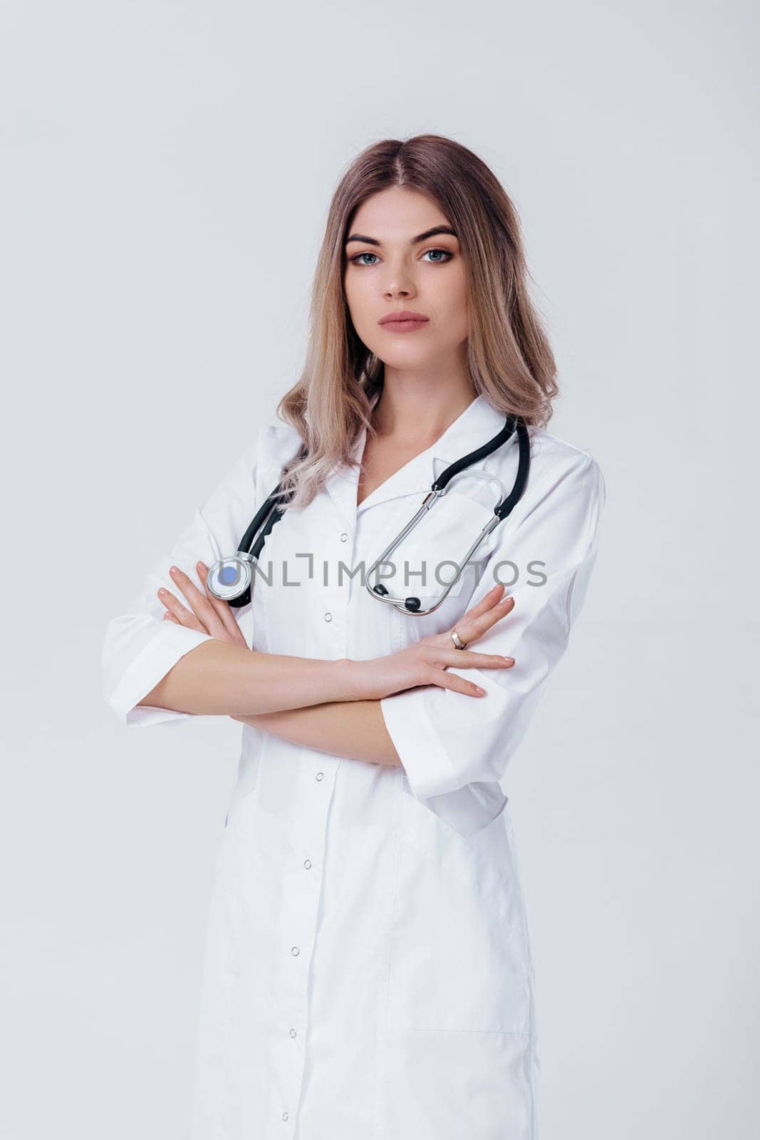 Medical physician doctor woman with crossed hands in white coat with stethoscope looks at camera on light background.
