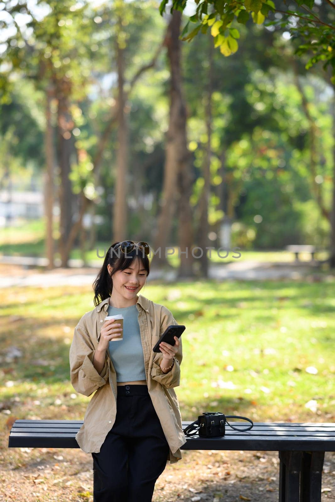 Portrait of pretty Asian woman in casual clothes holding paper cup of coffee and using mobile phone in the park by prathanchorruangsak
