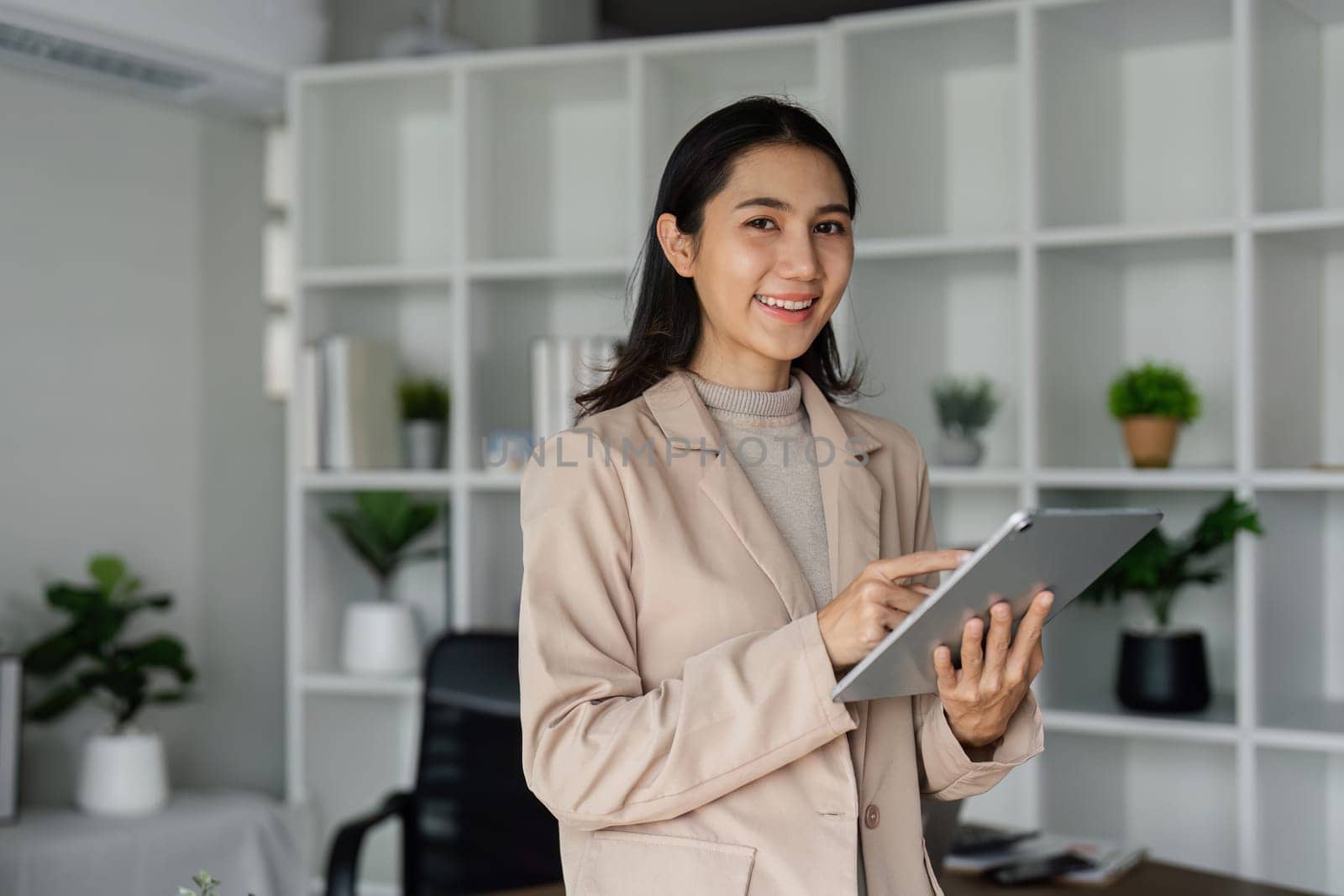 young asian woman, company worker in suit, smiling and holding digital tablet, standing over working space background.