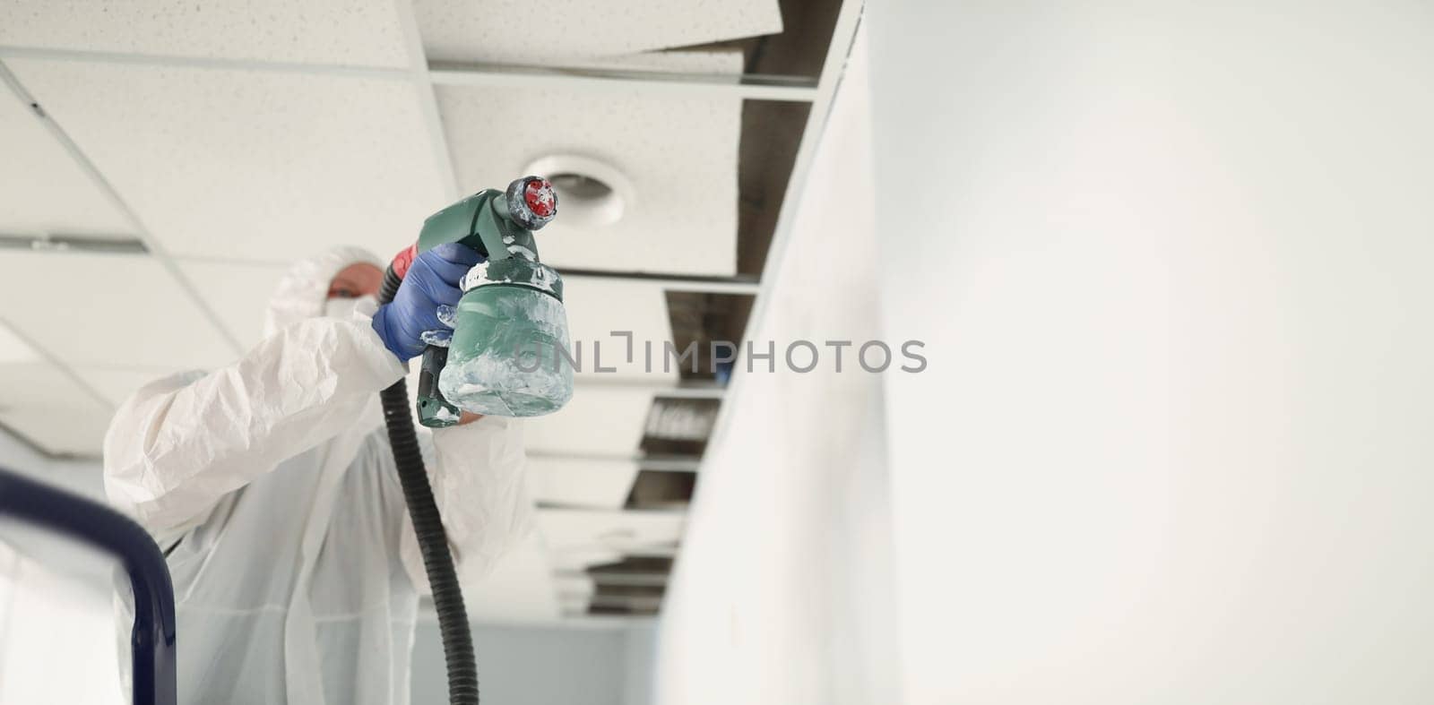 Male construction worker in protective overalls painting wall white with spray gun closeup. Finishing of apartments concept