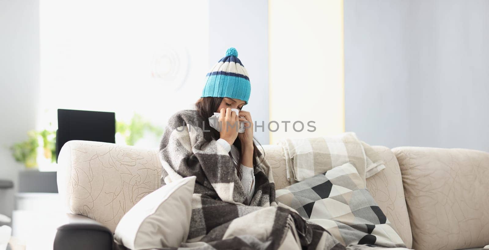 Sick woman in warm hat blowing her nose into paper napkin at home. Treatment of seasonal colds at home concept