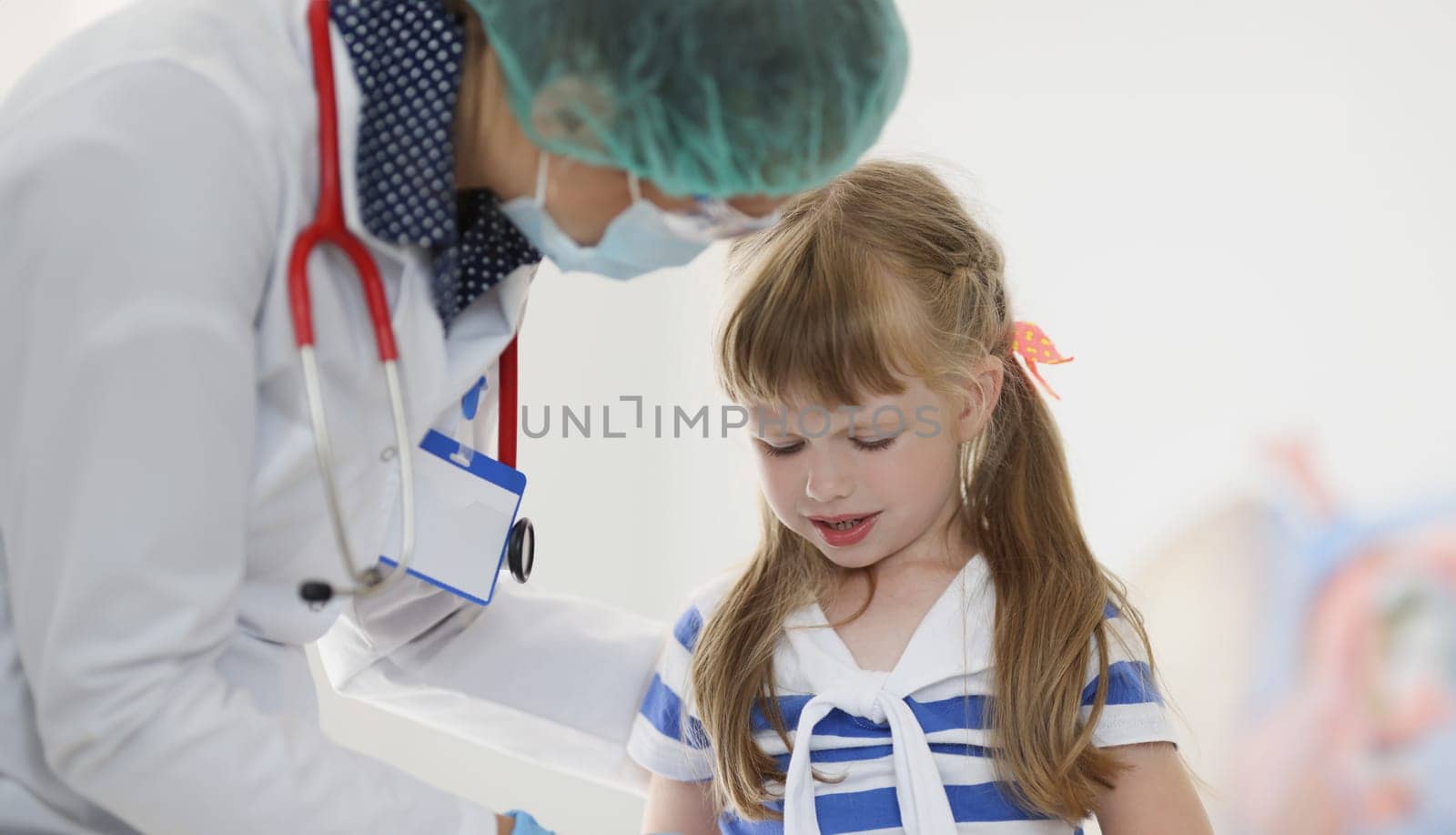 Doctor pediatrician in protective medical mask talking to little girl patient in clinic. Treatment of covid19 in children concept