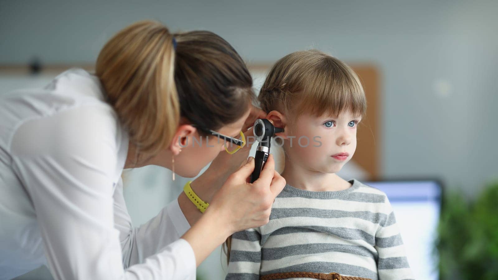 Female pediatrician looking at ear of little girl using otoscope in clinic by kuprevich