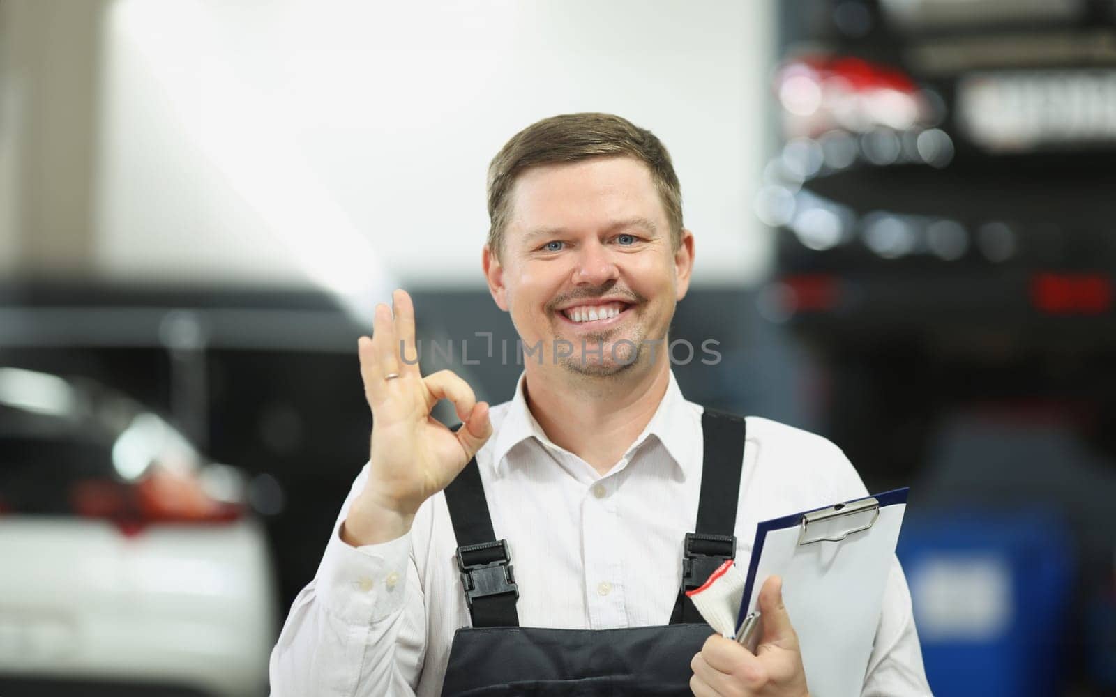 Master repairman holding documents and showing ok gesture in workshop. High quality diagnostics of car breakdown concept