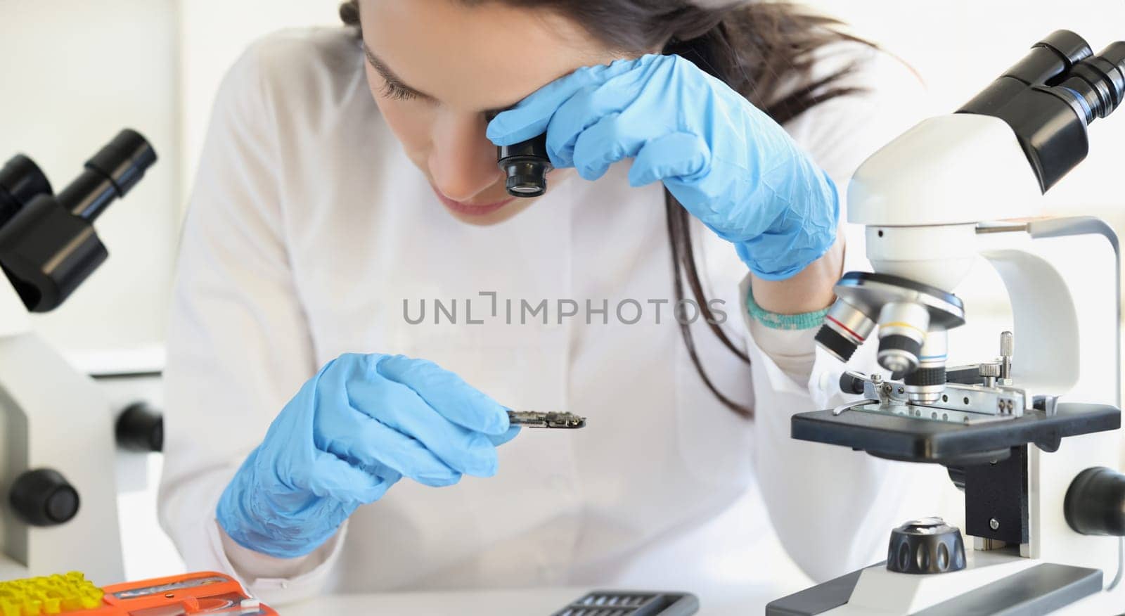 Woman scientist looking through magnifying glass at computer chip near microscope in laboratory. Innovative scientific inventions in electronics concept