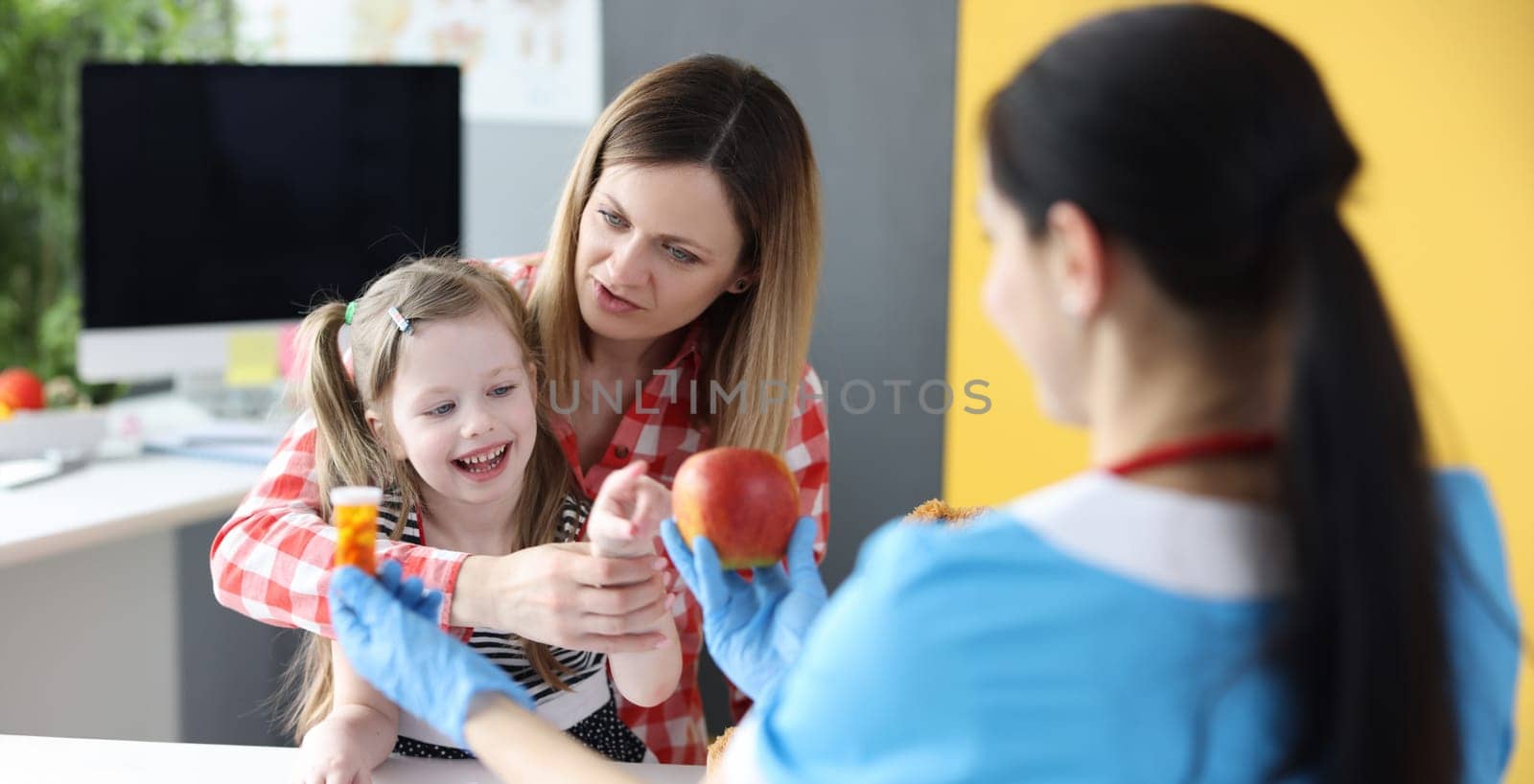 Pediatrician giving little girls choice of fruit apple or jar of vitamins in clinic by kuprevich