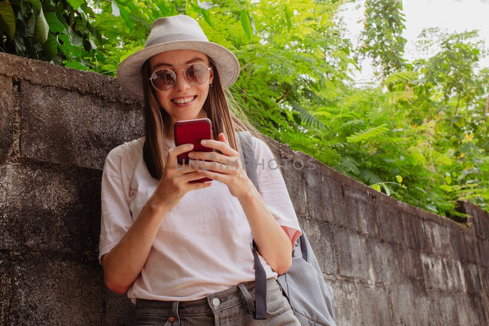 Beautiful young laughing lady in eyeglasses, hat and t-shirt looking at camera near brick wall and green bushes