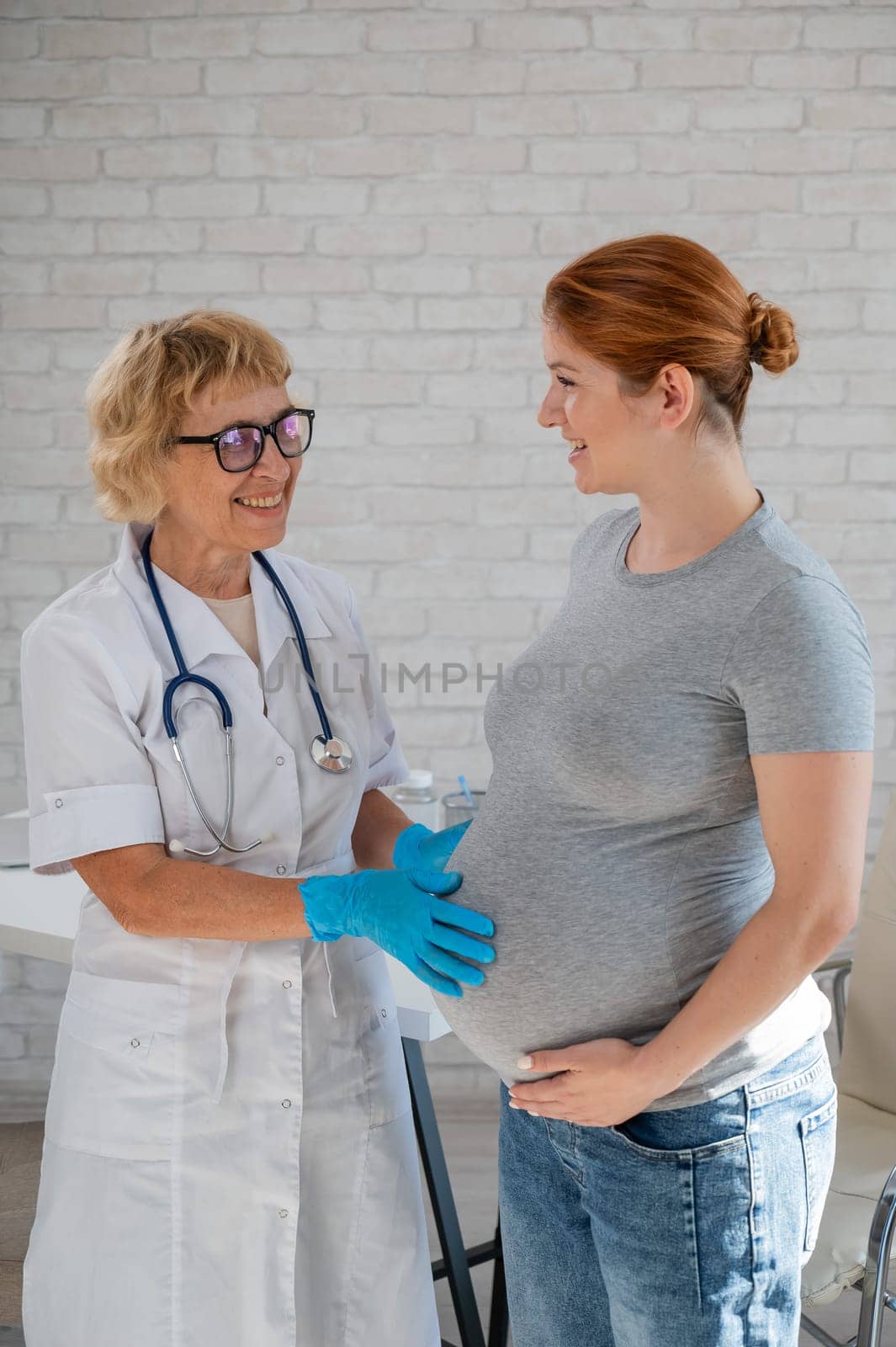 Pregnant woman visiting a doctor. Elderly Caucasian female gynecologist holds hands on the tummy of a pregnant patient. Vertical photo. by mrwed54