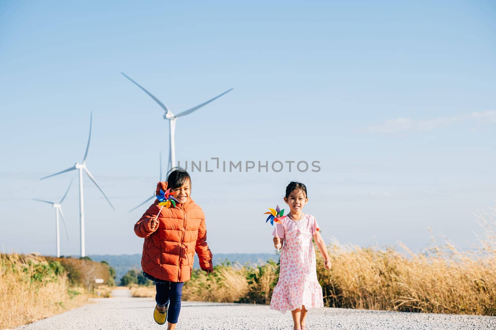 Two kids girl holding pinwheel joyfully run by windmills. Family near turbines embodies clean energy essence. Children happiness windmill circle against sun symbolizes sustainable cheerful community.