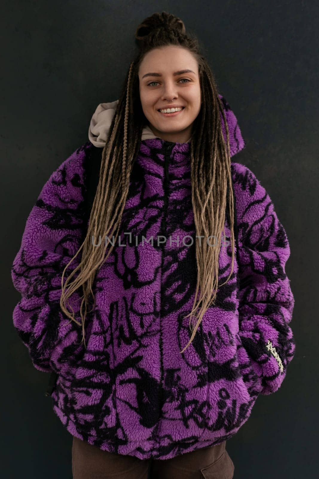 Stylish informal woman with dreadlocks and piercings is dressed in a purple jacket against the background of the street.