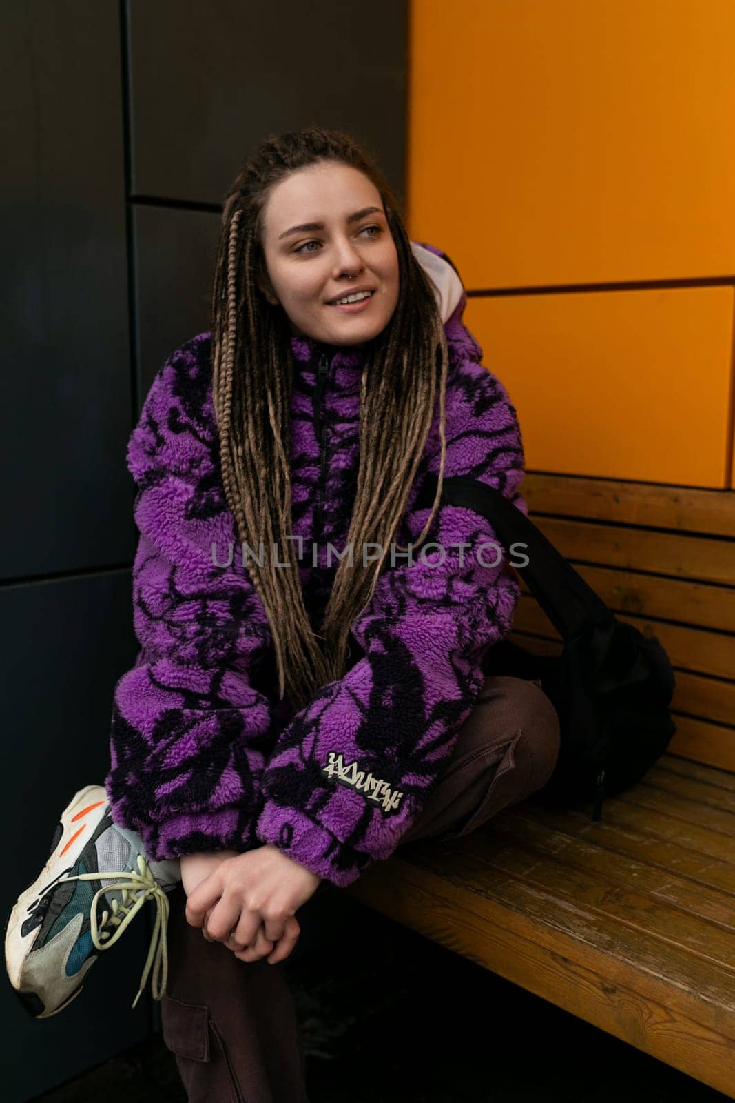 Stylish informal woman with dreadlocks dressed in a purple jacket walks through the city in winter by TRMK