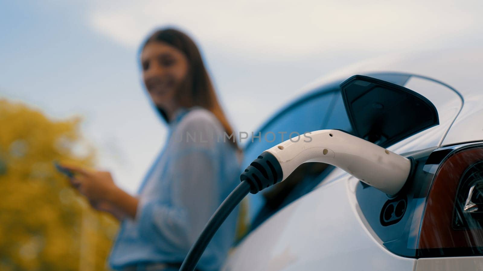 Woman recharging battery for electric car during road trip travel EV car in autumanl season natural forest or national park. Eco friendly travel during vacation and holiday. Exalt