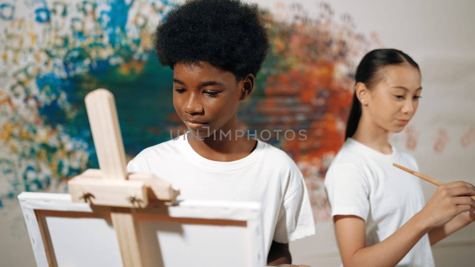 African boy painting canvas with watercolor while happy caucasian girl draw cool tone picture at colorful stained wall. Multicultural highschool student attend creative activity together. Edification.