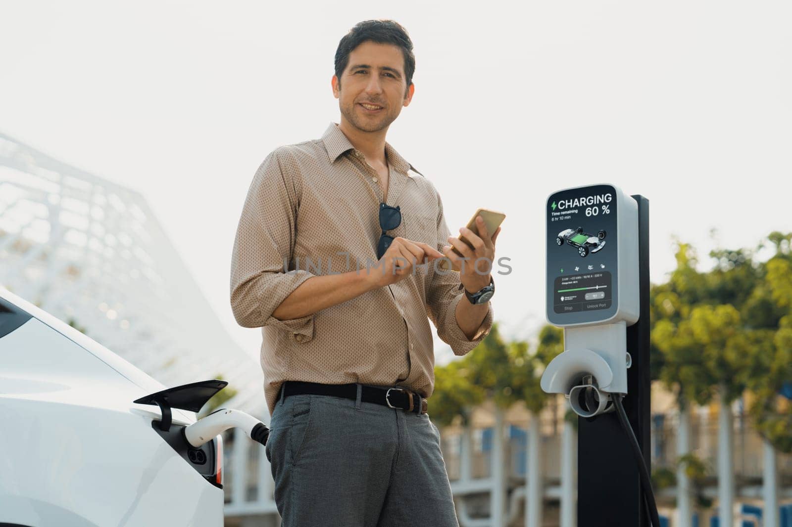 Young man use smartphone to pay for electricity at public EV car charging station green city park. Modern environmental and sustainable urban lifestyle with EV vehicle. Expedient