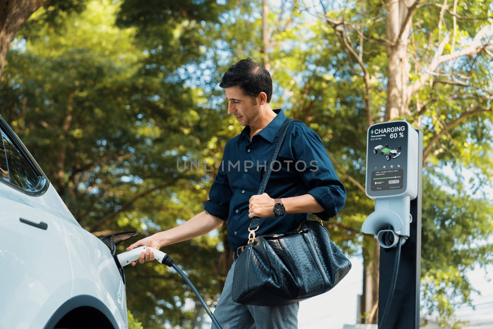 Young man recharge electric car's battery from charging station in outdoor green city park in springtime. Rechargeable EV car for sustainable environmental friendly urban travel lifestyle. Expedient