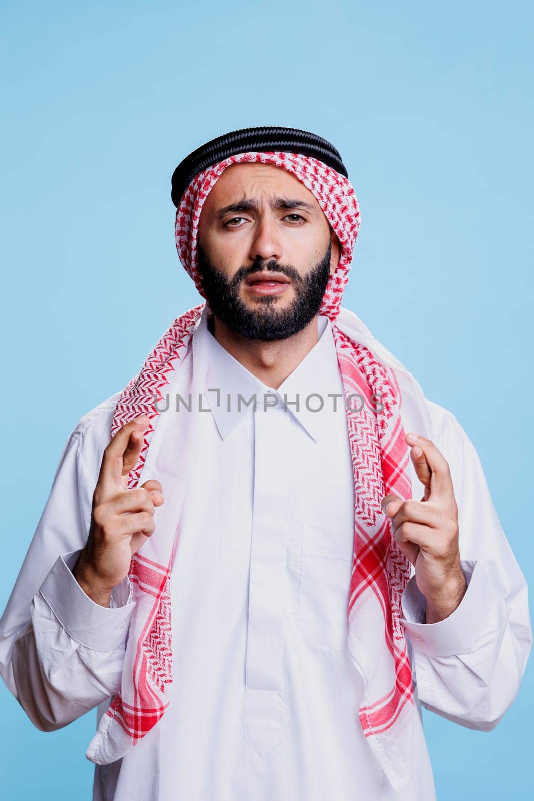 Muslim man dressed in traditional clothes making wish with crossed fingers and looking at camera with hopeful expression. Arab wearing thobe praying for luck with superstitious gesture portrait