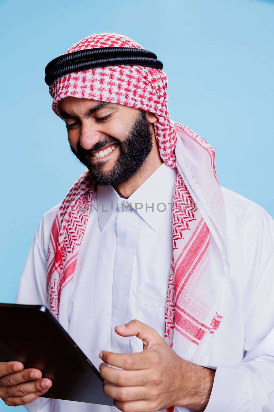 Arab man laughing while enjoying internet entertainment activity on digital tablet. Smiling muslim person having fun while chatting online and scrolling social media on gadget touchscreen