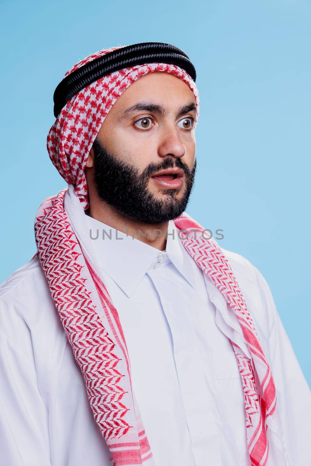 Shocked muslim man dressed in traditional headscarf looking away with wide eyes and open mouth. Person wearing arab ghutra and thobe clothes while posing with surprised face expression