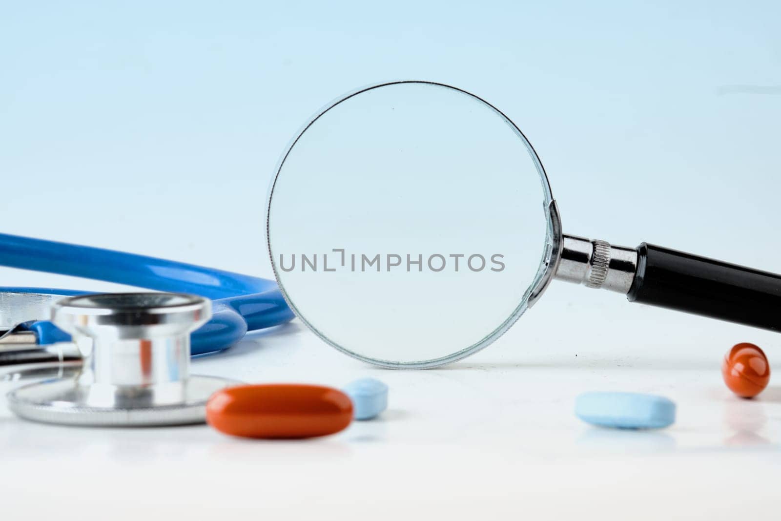 A magnifying glass on a blue background with pills in the foreground and a stethoscope. A conceptual photo. A place to copy.