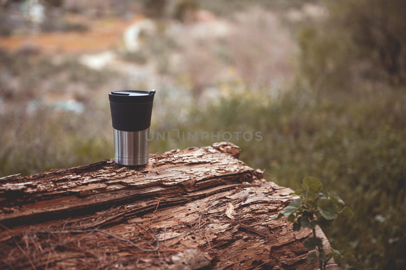 Still life with a stainless steel travel thermos mug on the log over beautiful early spring nature background. A mug of hot tea or coffee drink for enjoying scenery in nature. Trekking and wanderlust