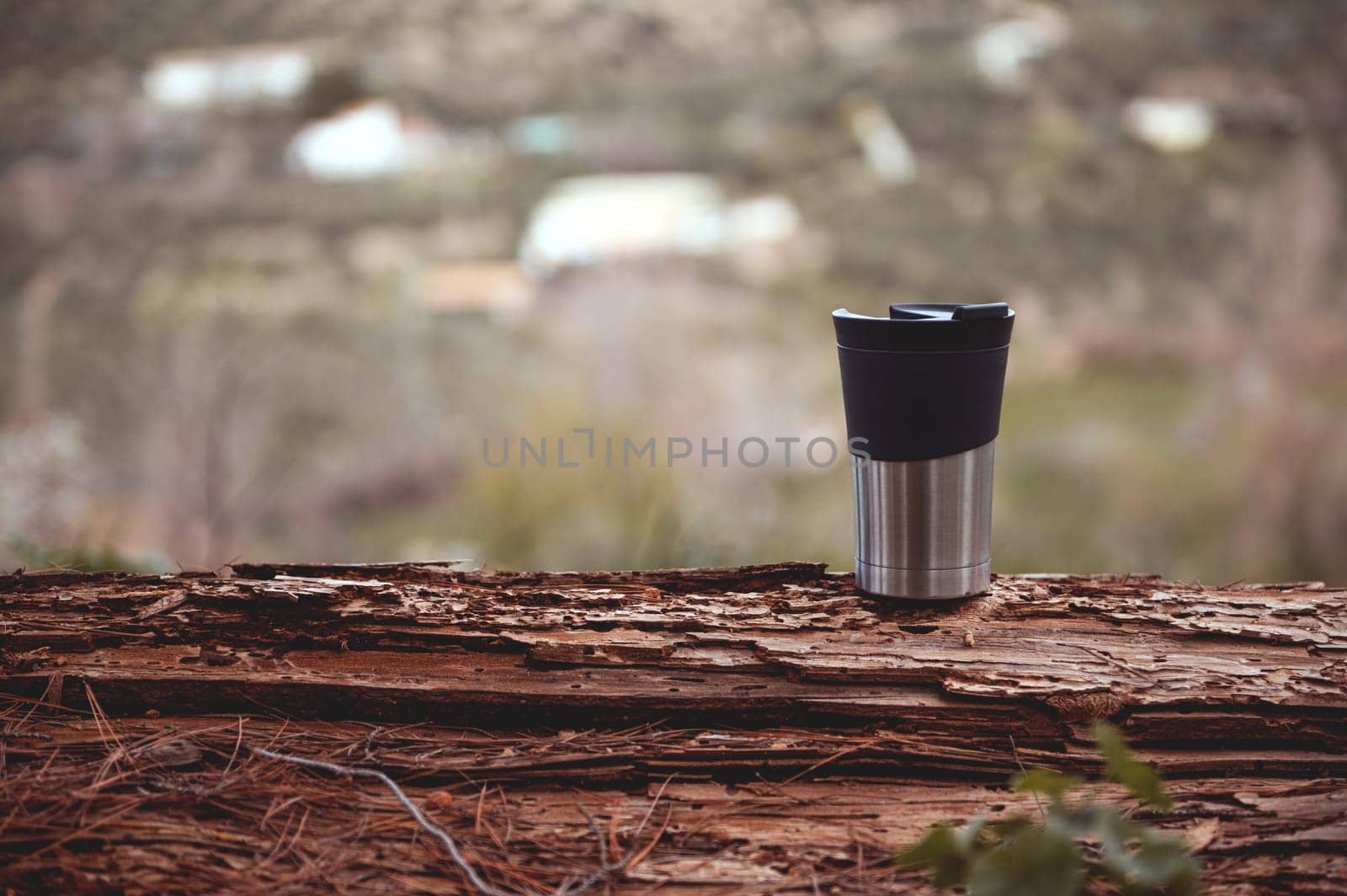 Horizontal shot of travel mug on a log over blur nature background with copy advertising space. A stainless steel cup with hot drink, coffee or tea for a recreation while trekking, hiking, travelling
