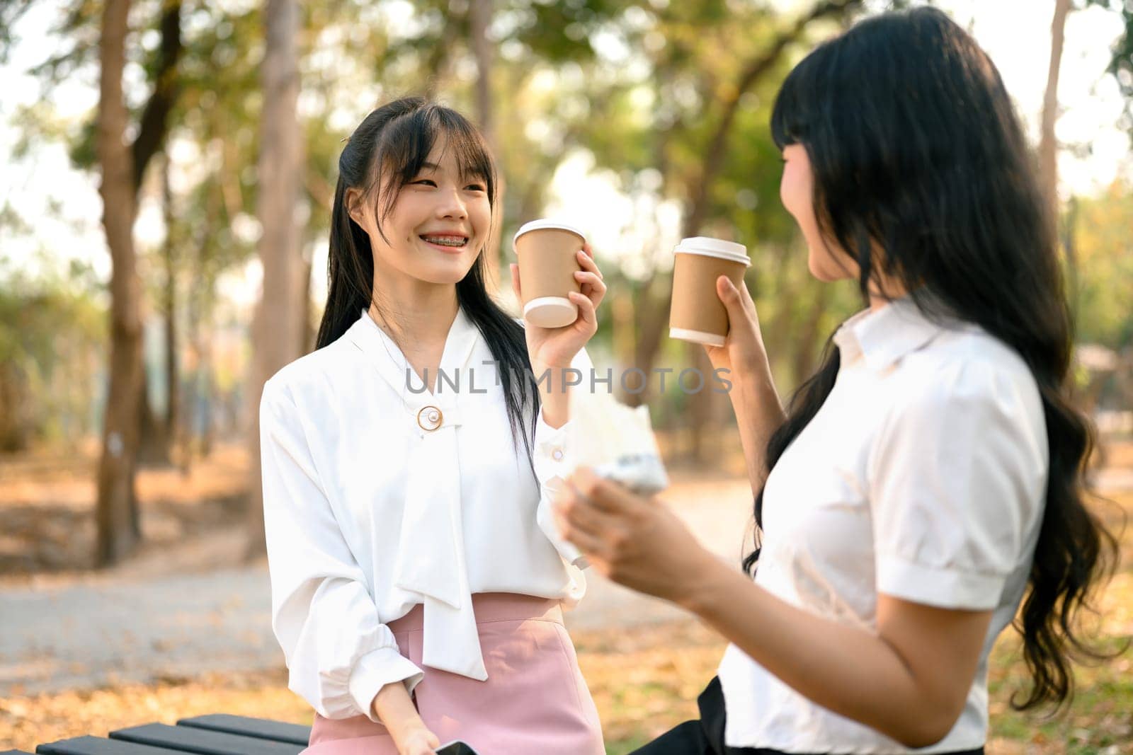Happy young Asian women drinking coffee from paper cups and chatting with each other in the park by prathanchorruangsak