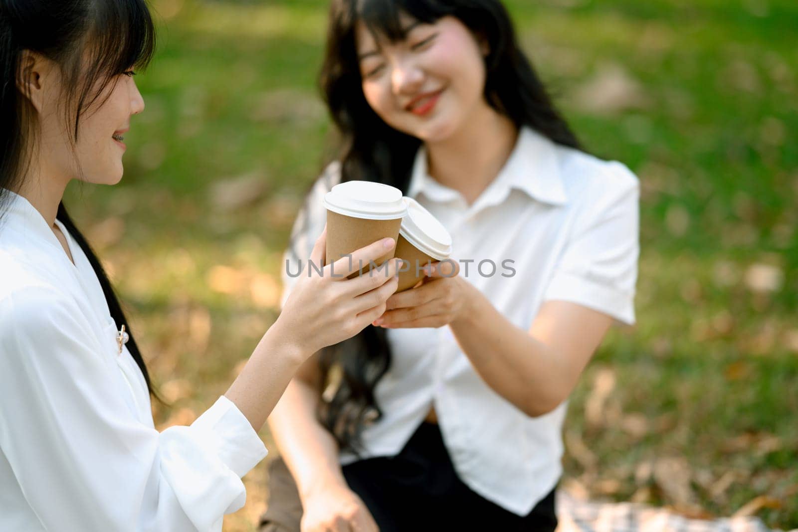 Smiling young woman clinking paper cups of coffee with her friend while relaxing together in the park by prathanchorruangsak