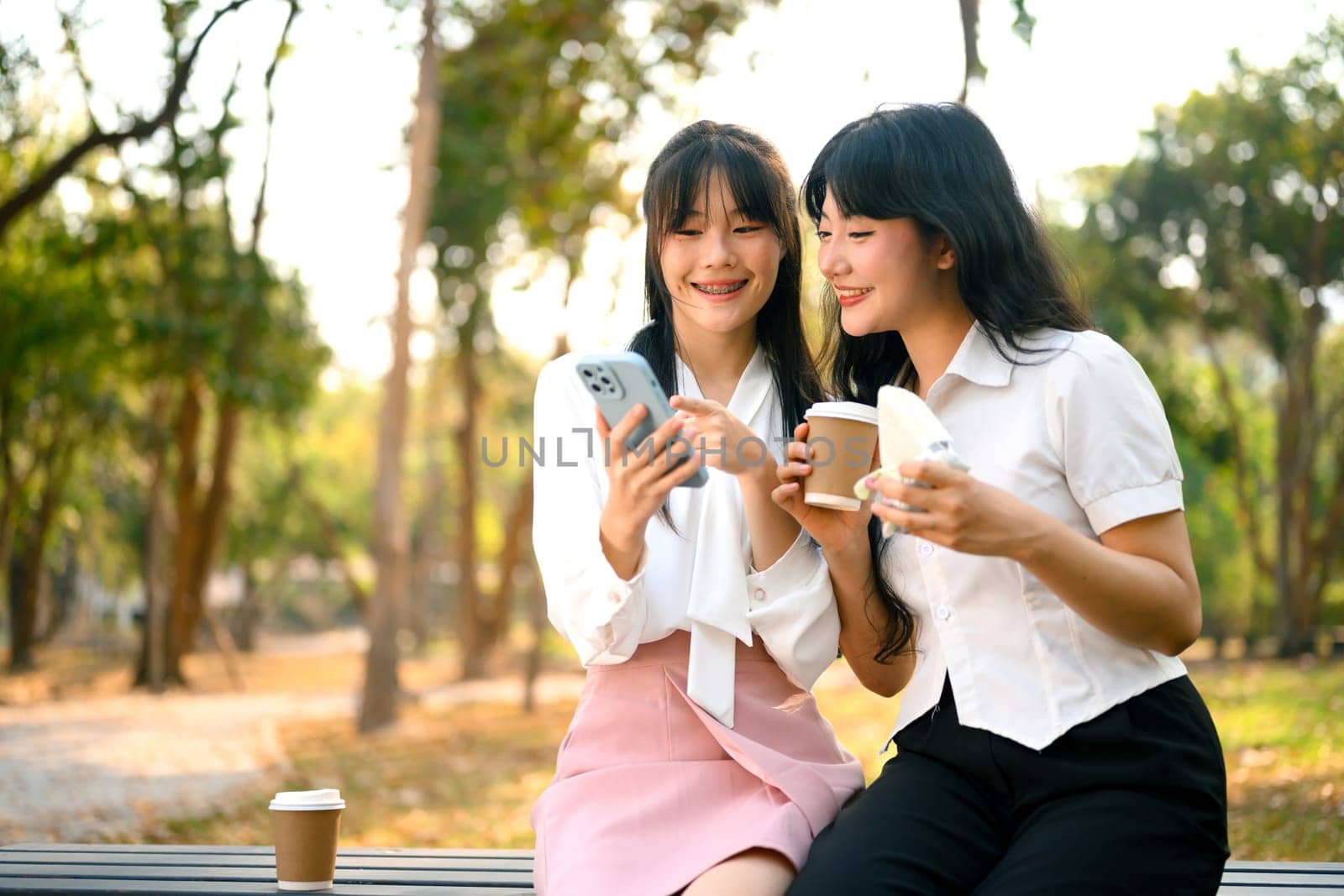 Two happy female friends talking and using mobile phone together in the the park by prathanchorruangsak