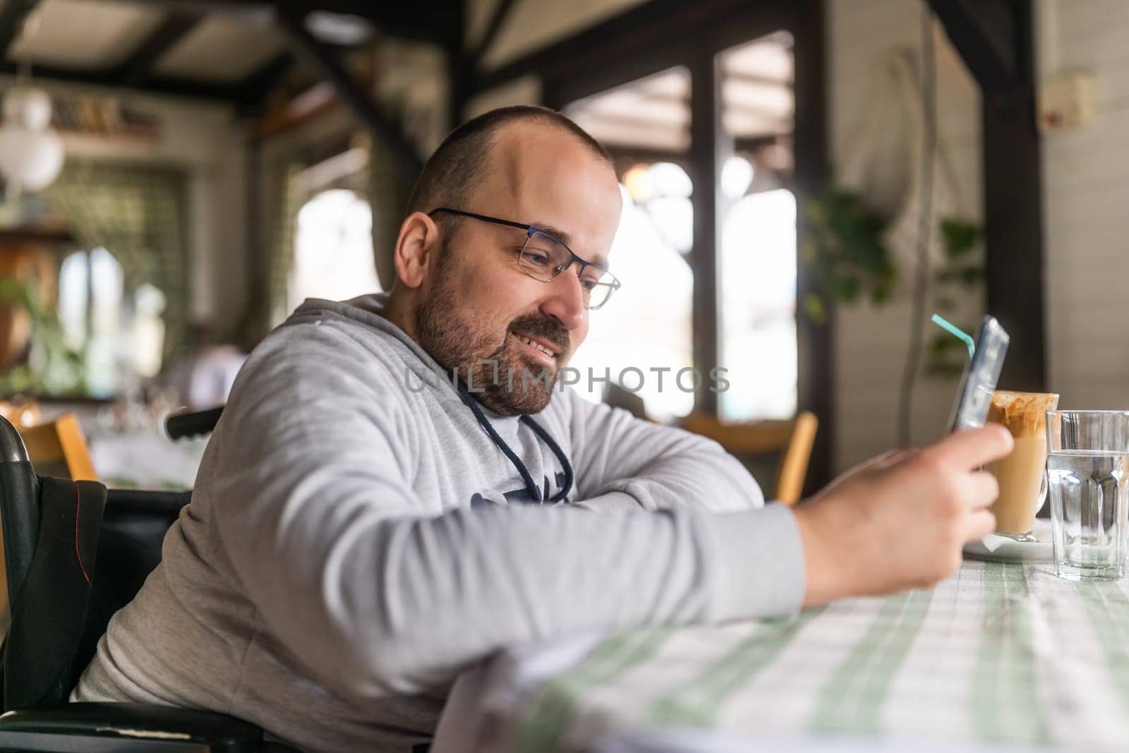 Happy paraplegic handicapped man in wheelchair is sitting at restaurant and using smartphone by djoronimo