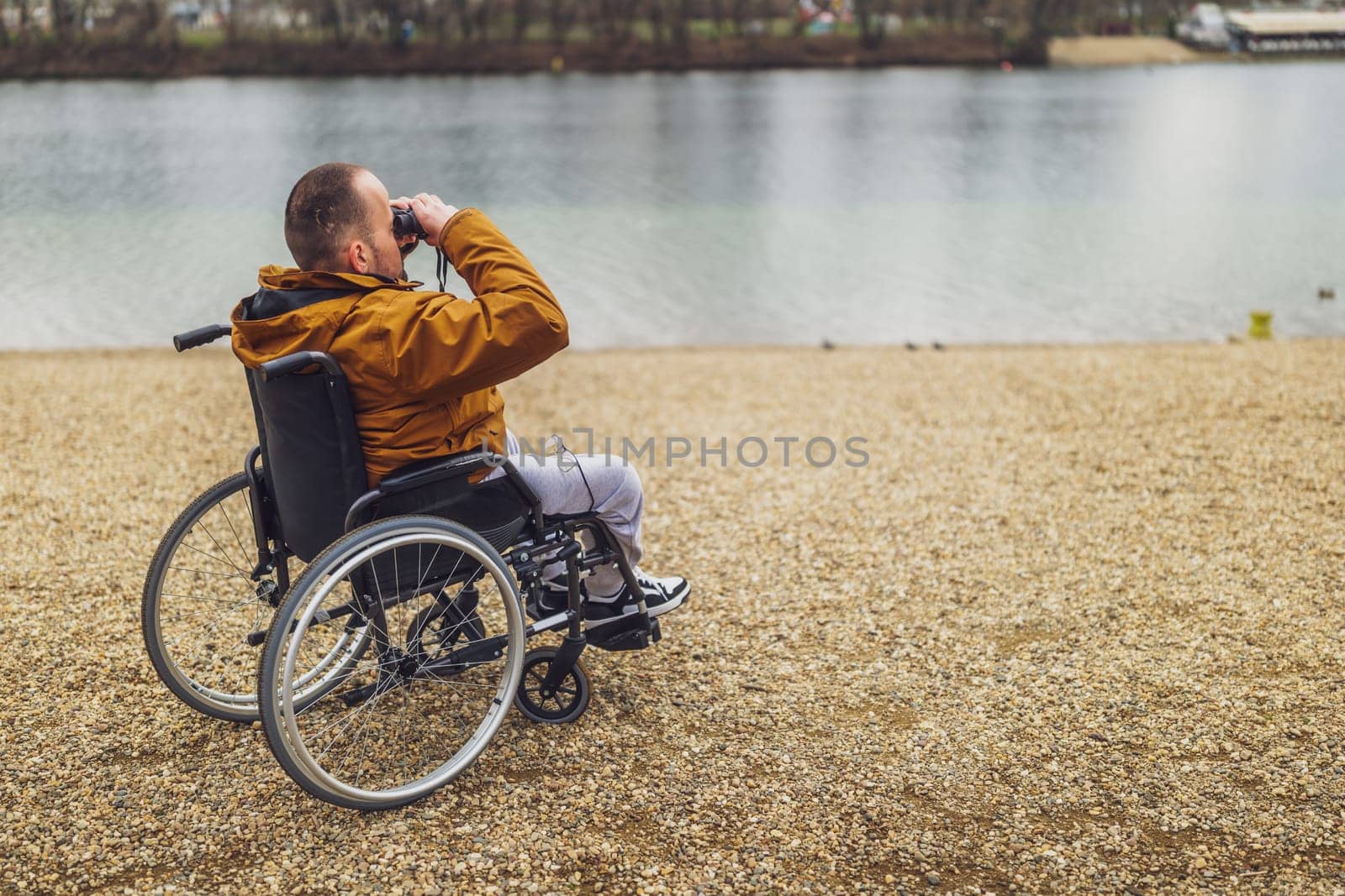 Paraplegic handicapped man in wheelchair is using binoculars outdoor. He is watching nature by the lake.