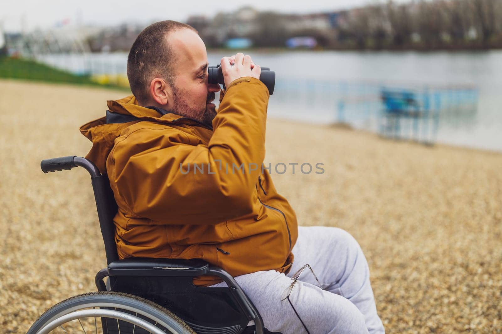 Paraplegic handicapped man in wheelchair is using binoculars outdoor. He is watching nature by the lake.