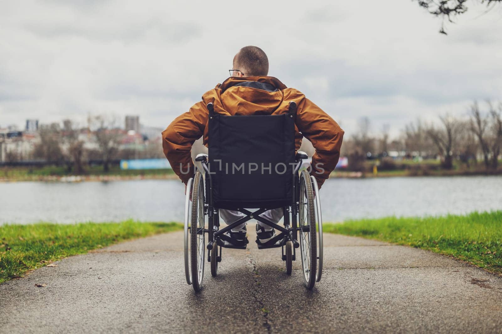 Rear view image of paraplegic handicapped man in wheelchair by the lake. He is rolling on walkway.