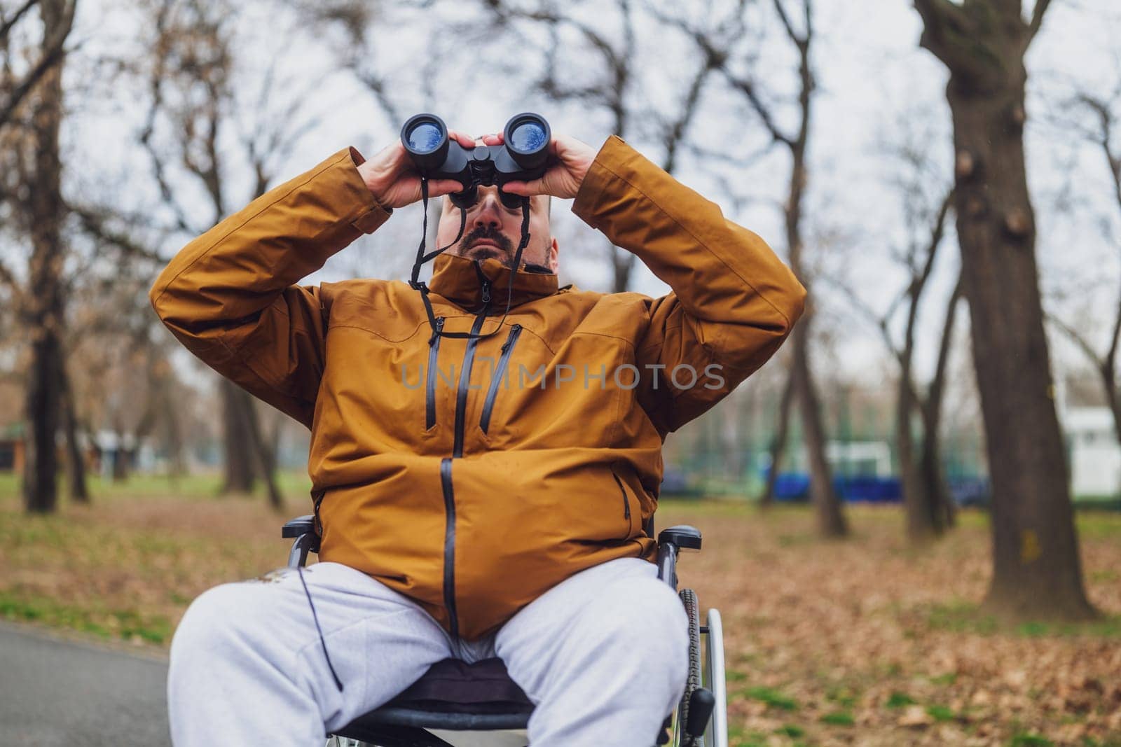 Paraplegic handicapped man in wheelchair is using binoculars outdoor. He is watching birds.