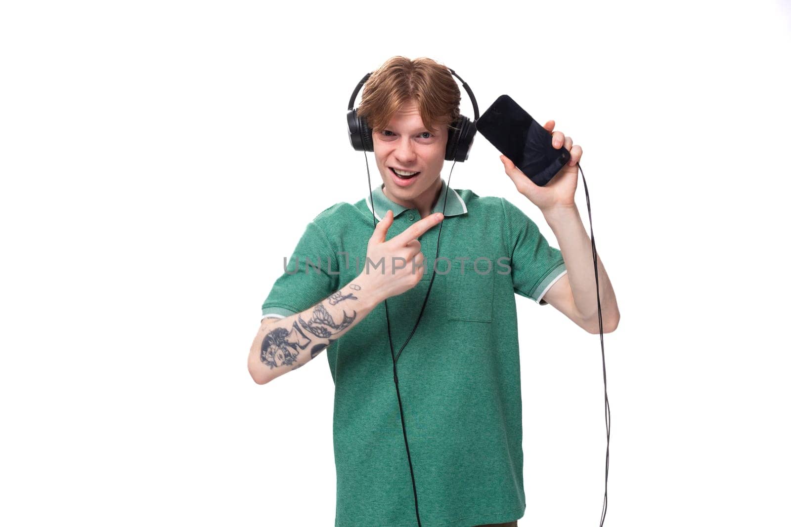 young cheerful slender student european man with red hair in a green t-shirt listens to loud music in headphones by TRMK